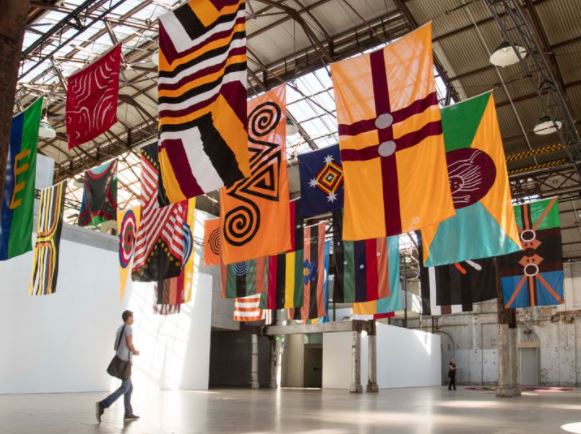 Archie Moore, United Neytions (2014–2017). Installation of 28 flags, polyester, nylon, zinc plated alloy. 28 parts in two sizes: 23 x 360 x 180 cm and 5x 180 x 180 cm. Exhibition view: The National: New Australian Art, Carriage works, Sydney (2017). Courtesy the artist and The Commercial, Sydney. Photo: Sofia Freeman.